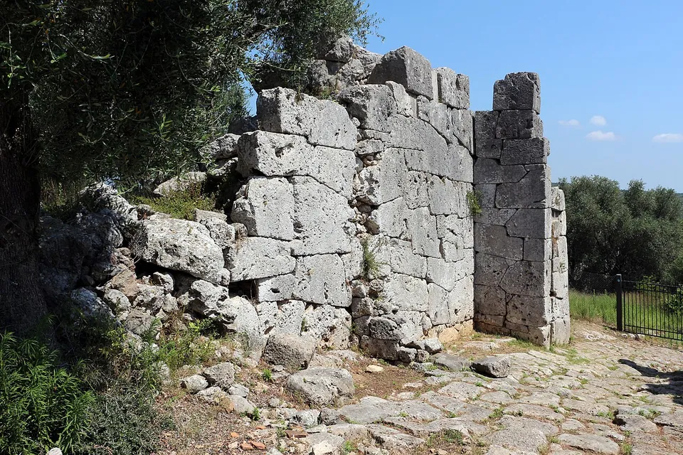 A tall pier and wall of large squared grey stone blocks beside a stretch of old paving, under an olive tree, with a modern iron fence beyond.