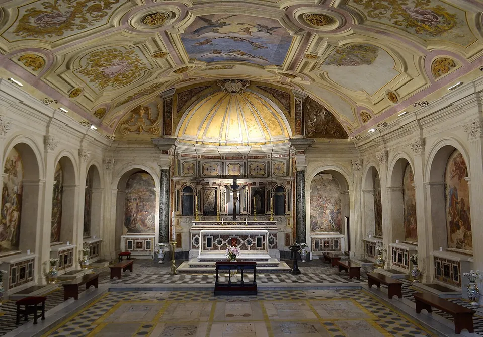 A low, wide crypt with a painted and gilded vault, side altars set in arches along both walls, a marble high altar under a small dome at the far end, and a patterned tiled floor.