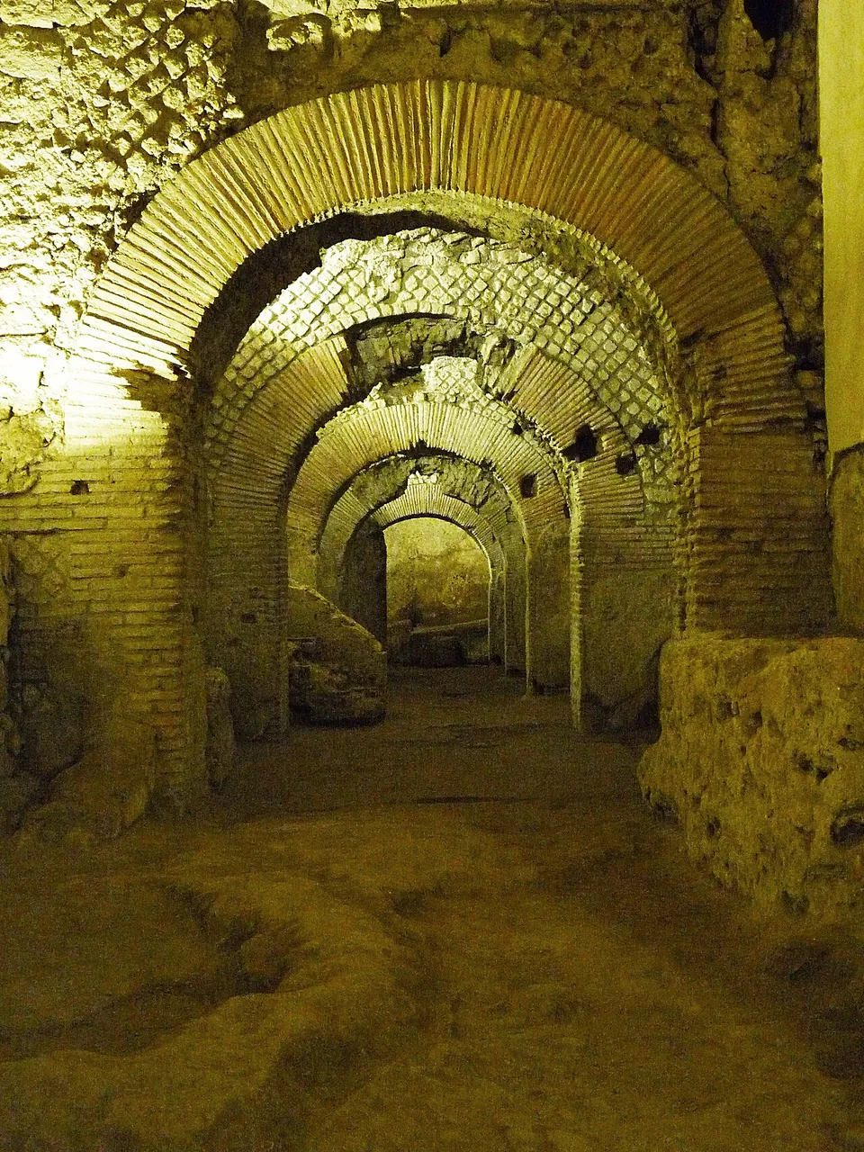 A row of brick arches recedes into the distance under a vaulted ceiling, lit yellow, with an uneven earth floor and broken masonry along the sides.