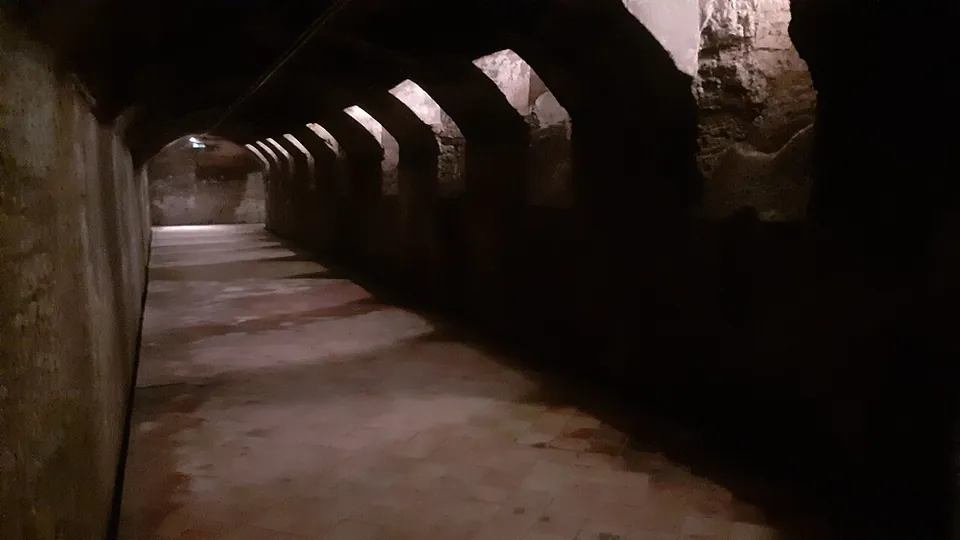 A long barrel-vaulted corridor underground, its floor of reddish tiles, with a row of small splayed openings high in the right-hand side of the vault letting in pale light.
