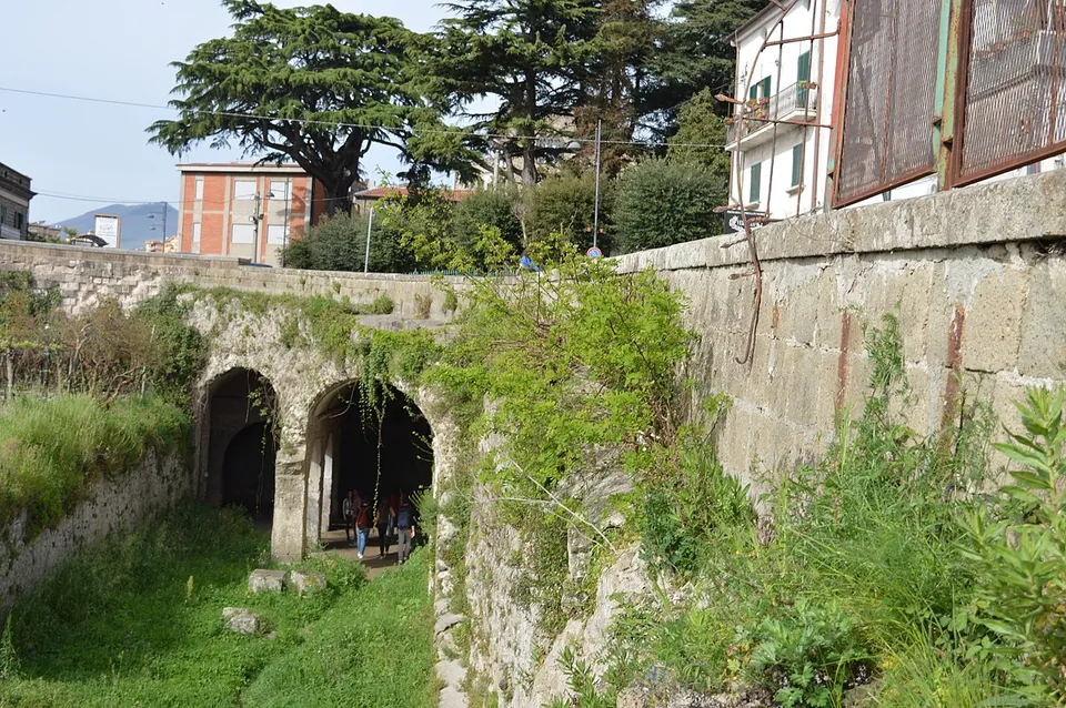 Two round-arched openings at the foot of a high stone retaining wall, reached across a grassy sunken strip, with visitors standing inside the shaded passage and modern houses and trees on the street level above.