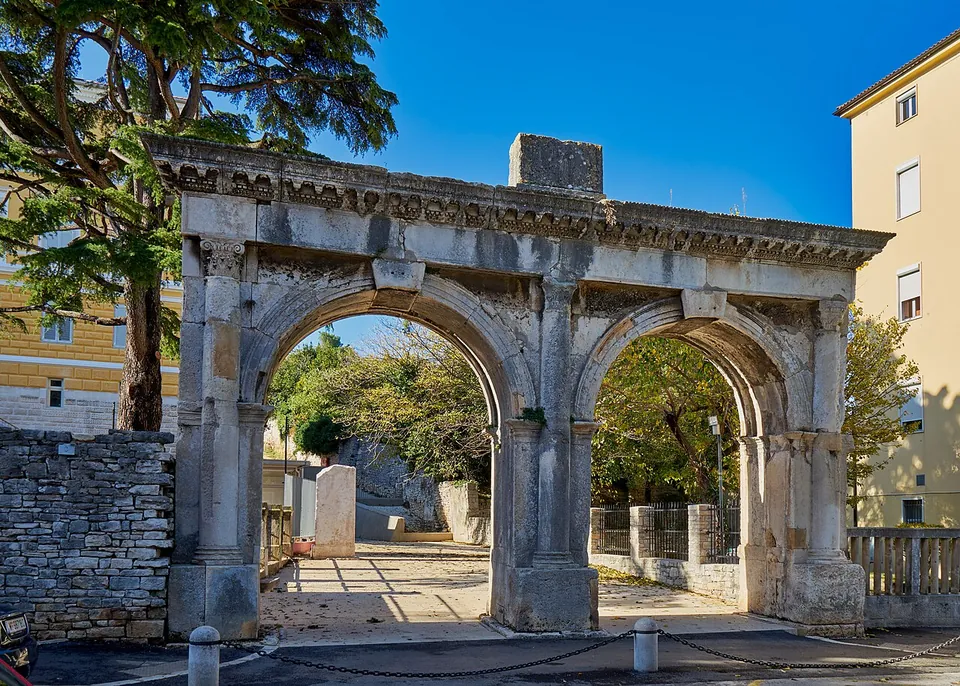 A Roman gateway with two equal round arches under one carved cornice, standing at the edge of a street with a stone wall on its left, trees behind and a paved path leading through.