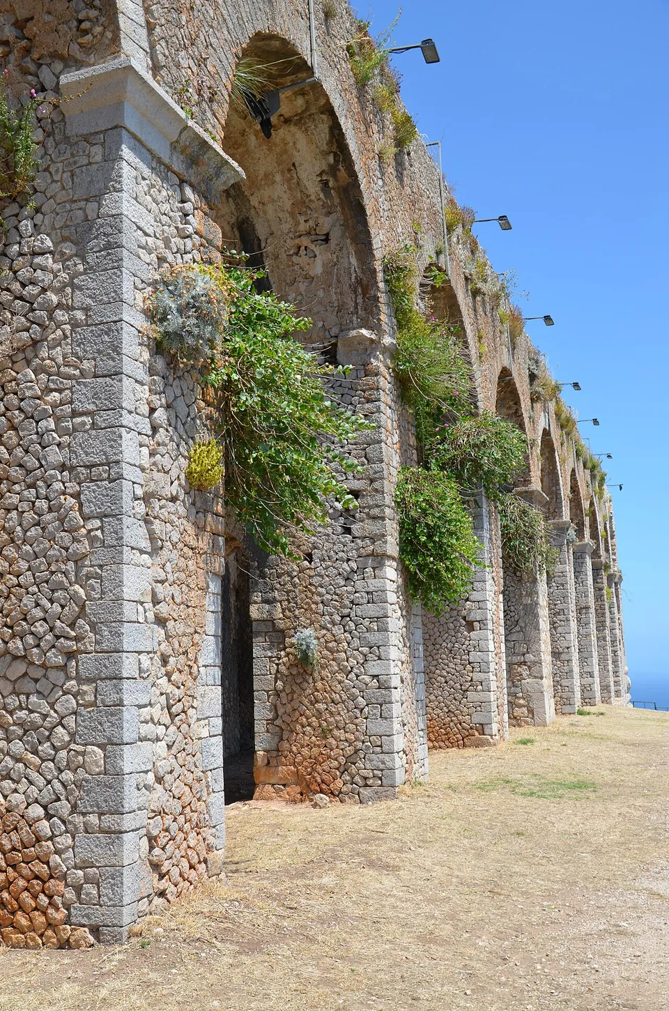 A row of tall round arches built of small rough stones with dressed blocks at the corners, plants hanging from the wall and lamps fixed along its top, receding along a dry gravel terrace under a blue sky.