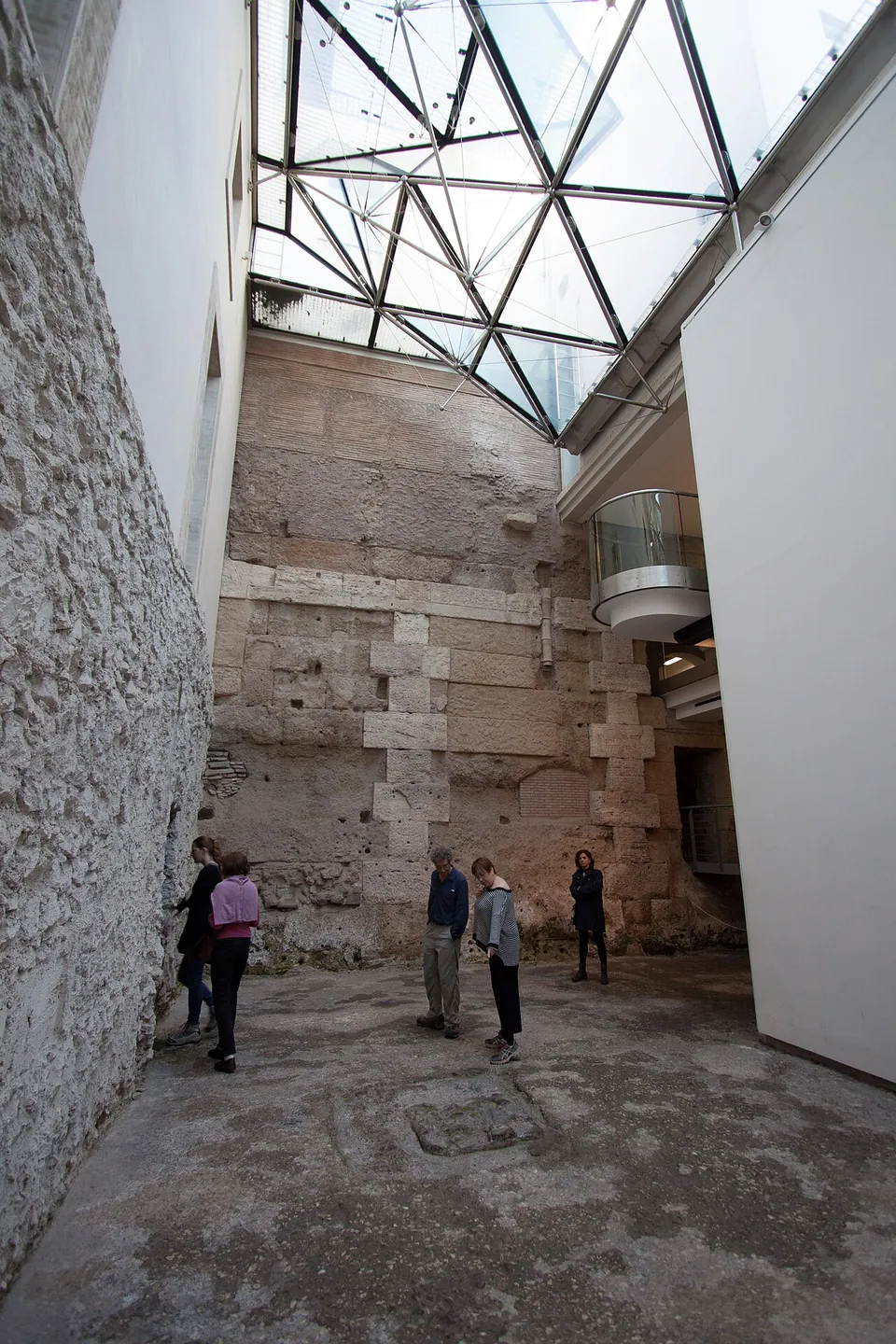 A tall courtyard inside the museum, walled on one side with rough masonry and at the back with squared stone blocks, lit from a faceted glass roof, with a few visitors standing on the uneven floor and modern white walls and a glass-fronted balcony at the edges.