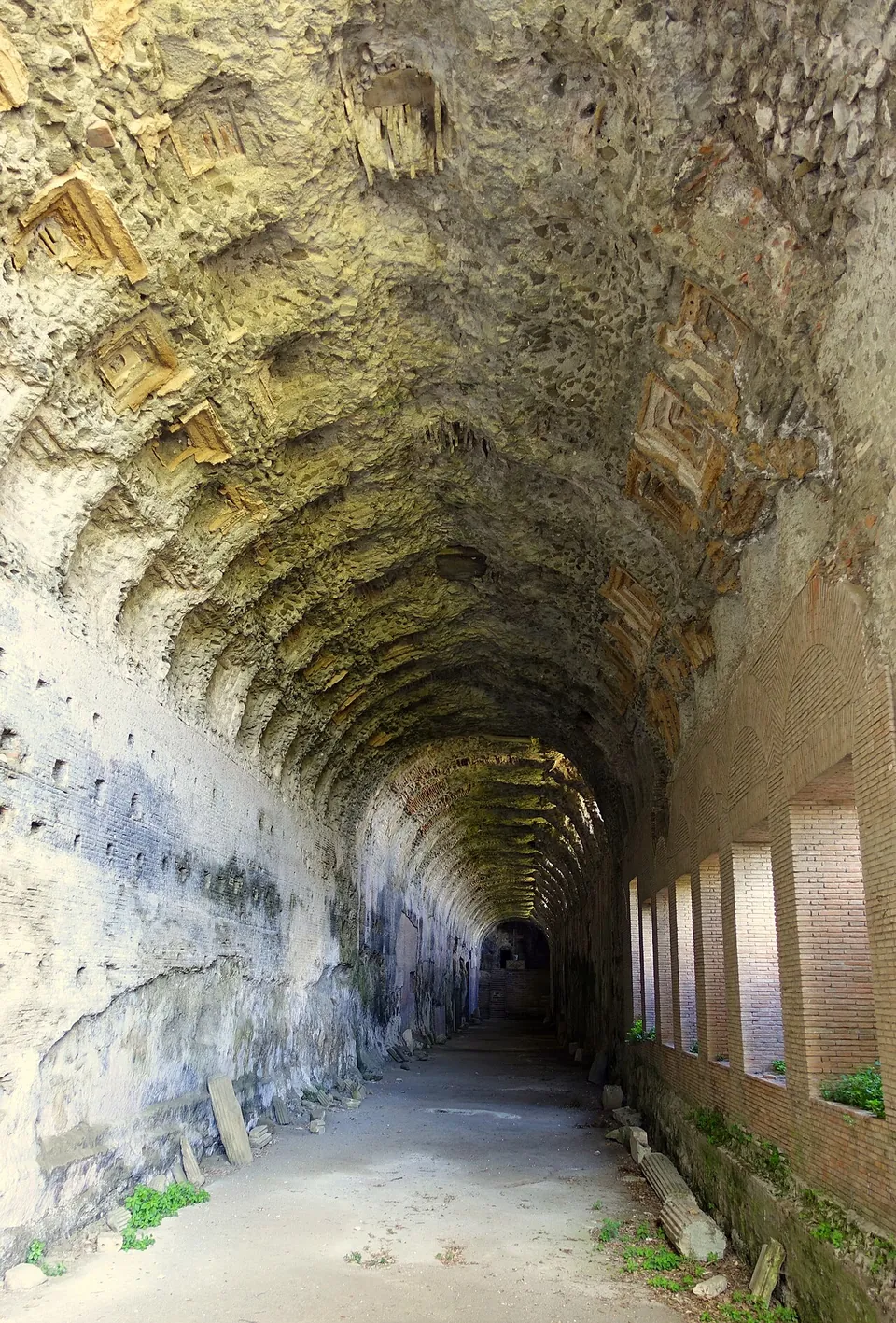 A long barrel-vaulted corridor of pale stone running into the distance. The vault is patterned with weathered square coffers, and a row of tall rectangular windows lets light in along the right-hand wall.