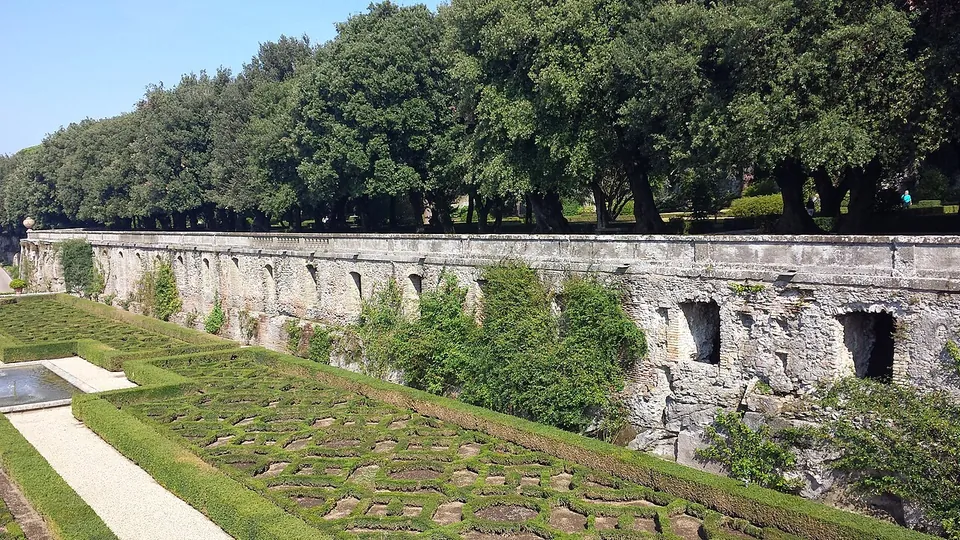 A long, weathered stone wall pierced by a row of rectangular openings. It rises behind a formal parterre of low clipped hedges, with a line of dense evergreen trees along its top.