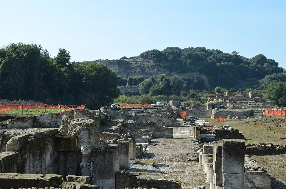 Excavated walls and a straight paved street running across the lower city, with orange safety fencing round open trenches and a wooded hill rising behind, with structures on its crest.