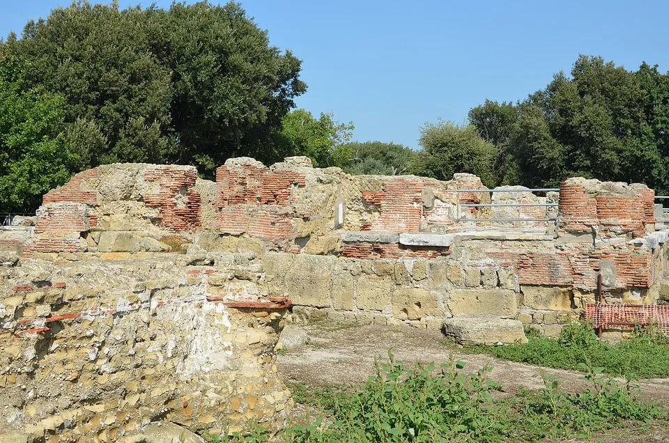 Low ruined walls of tufa blocks topped with courses of red brick, standing on a raised platform among weeds, with holm oaks behind.