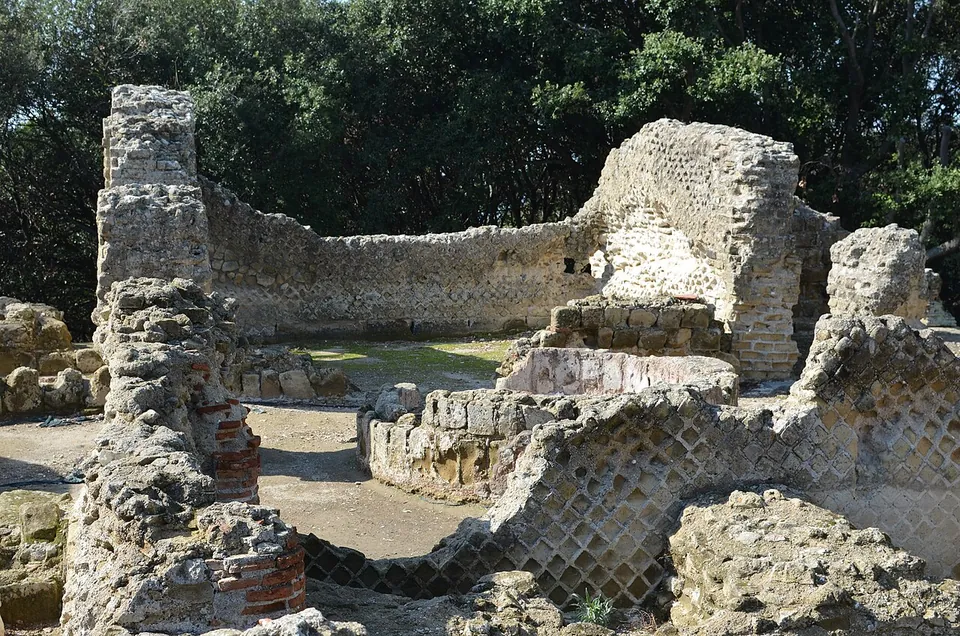 Ruined walls faced with a net-like pattern of small square stones, banded and patched with brick, enclosing an open interior where lower walls and a sunken structure stand among trees.