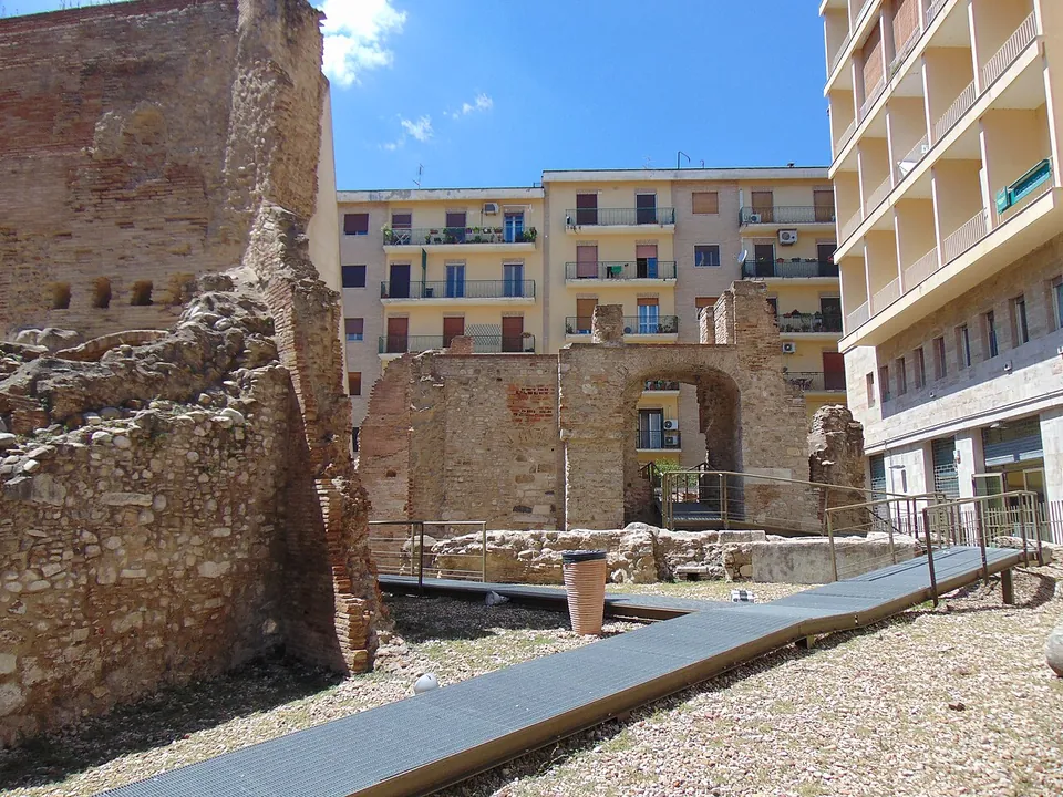 A raised metal walkway running across gravel between ruined walls of rubble stone and brick, towards a brick wall pierced by an arched opening, with blocks of flats behind.