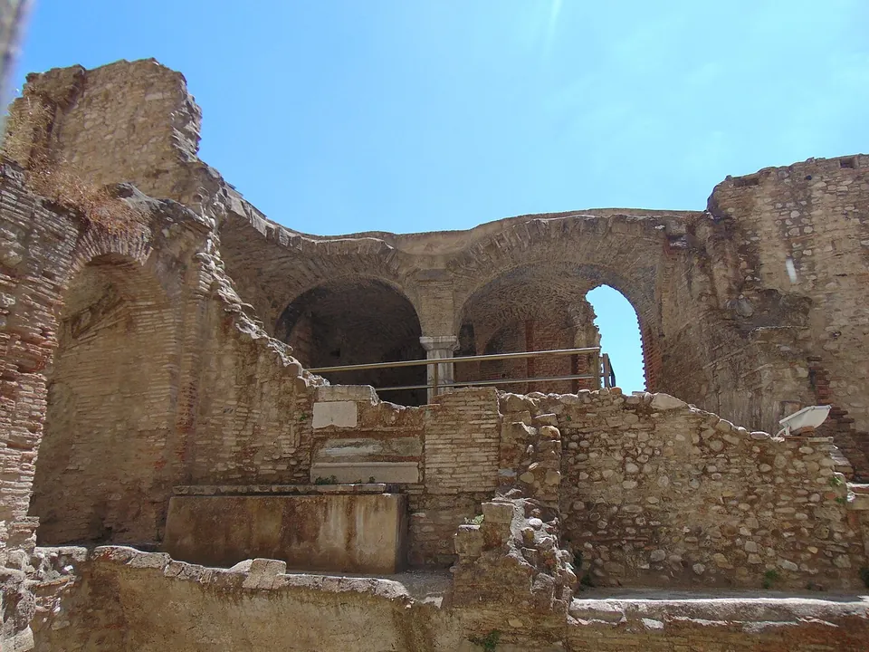 Ruined walls of brick and rubble stone standing to different heights, with a row of brick arches on piers at the back, seen from low down inside the excavation under a clear sky.