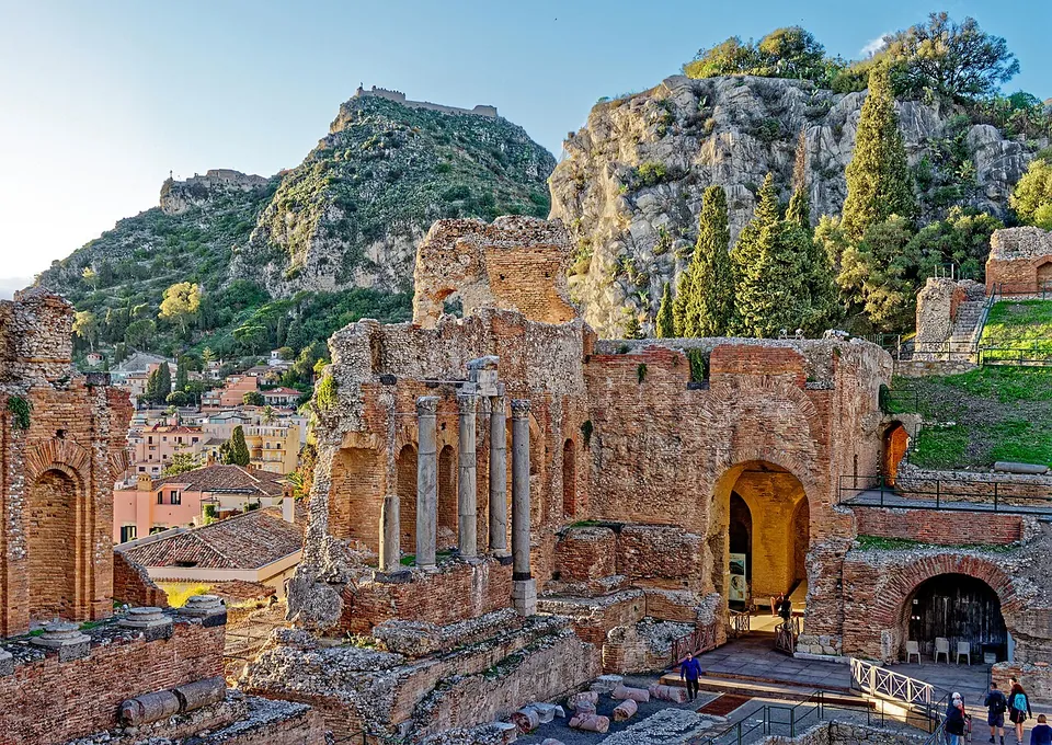 The ruined red-brick back wall of the stage, with round-headed niches and arches and a few tall stone columns standing in front of it, below a rocky hill crowned by ruined walls.