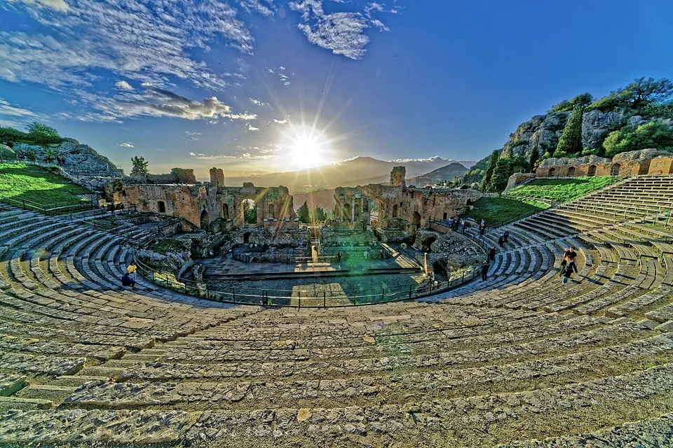 The view from high in the seating bowl down over curving stone tiers to the orchestra and the ruined stage building, with the sun setting over mountains beyond.