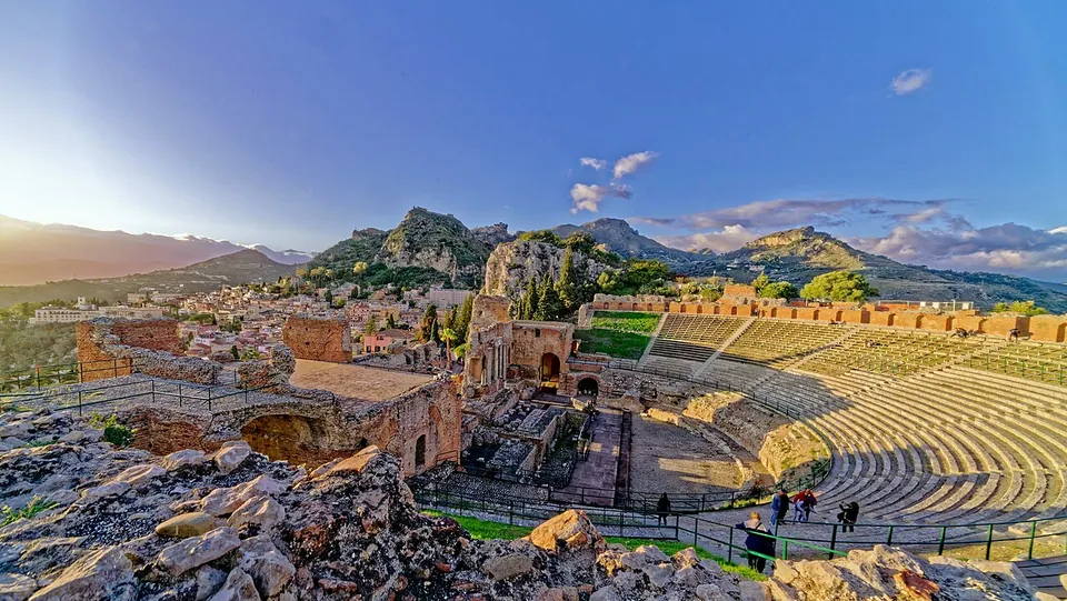 The theatre seen from rocks above one end, with the brick stage building in the middle distance, the curve of the seating to the right, and the town and a ring of mountains beyond under an evening sky.