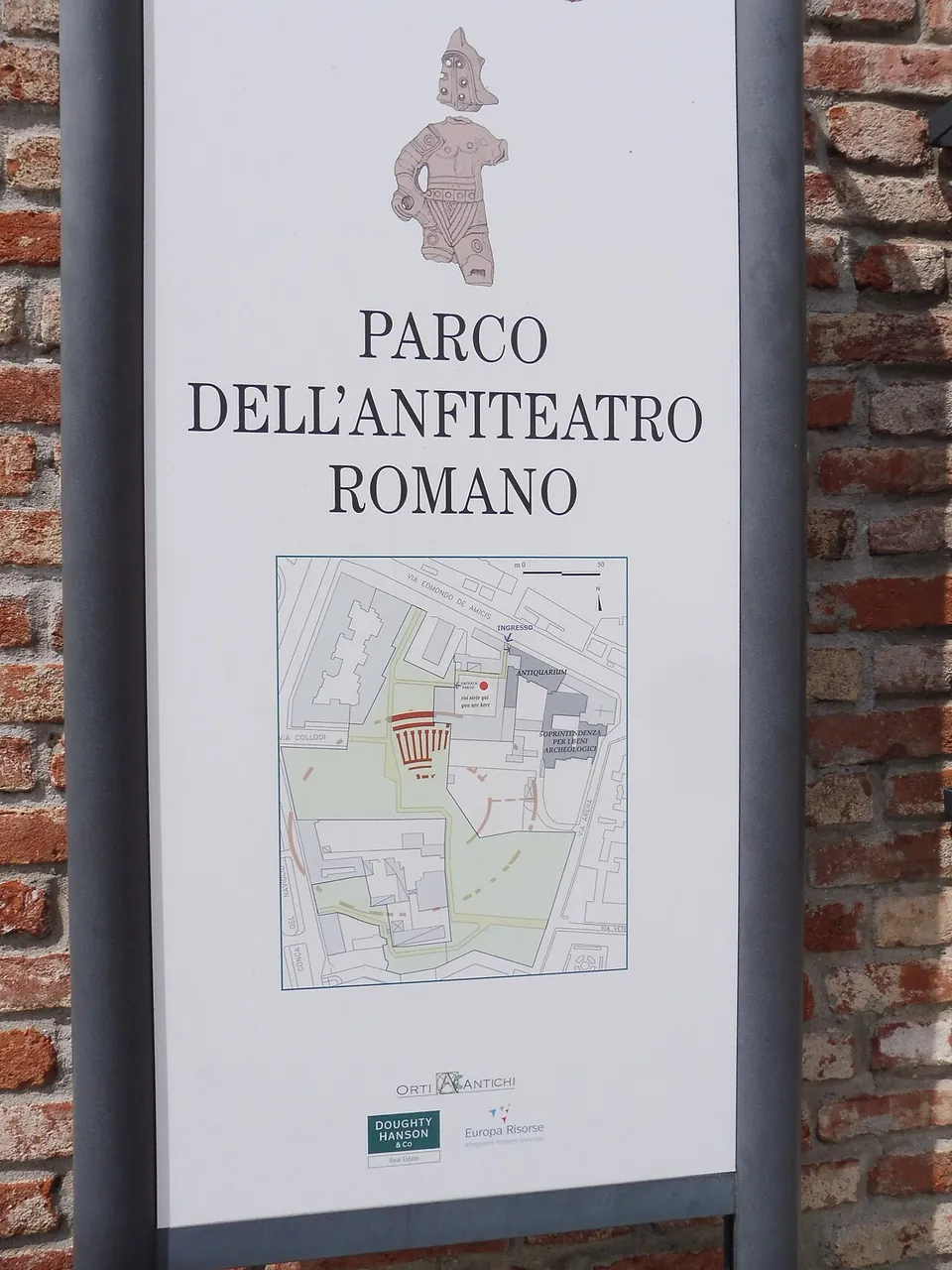 A white sign on a brick wall headed "Parco dell'Anfiteatro romano", with a drawing of a figure above the title and a plan of the park below it.