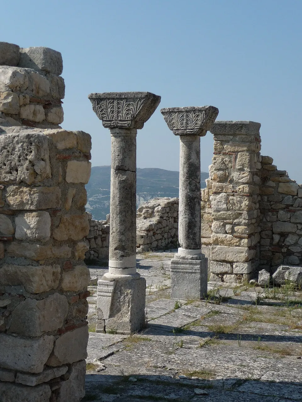Two stone columns with carved capitals standing on a paved floor between stone walls, with hills in the distance behind.