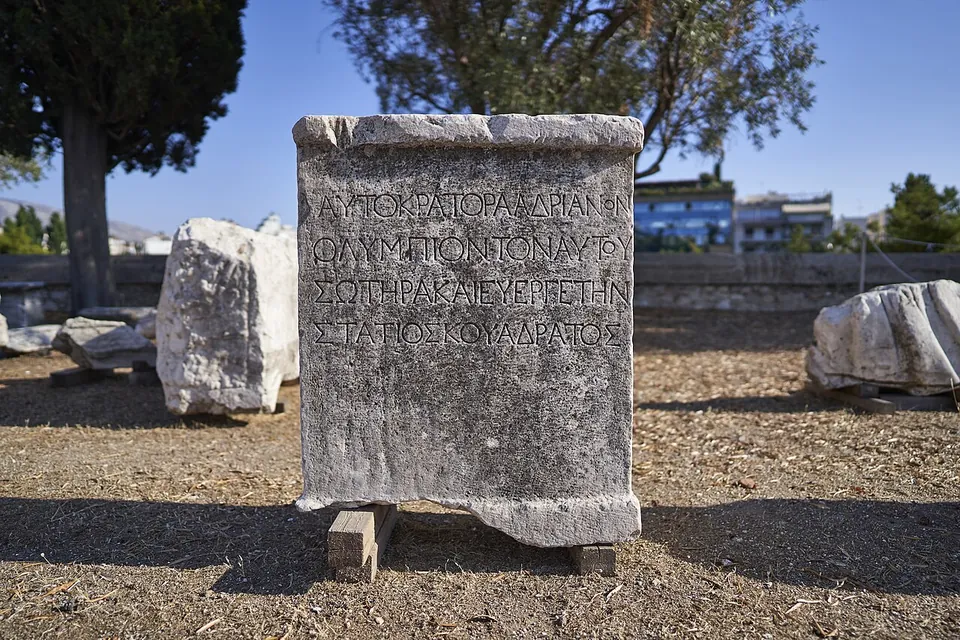 A weathered marble block standing upright on the ground, its face carved with four lines of Greek capitals, among other fallen blocks under trees.