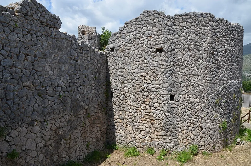 A stretch of wall and a round tower, both built of irregular rough-faced stones, the tower pierced by small square openings, standing on dry grass under a cloudy sky.