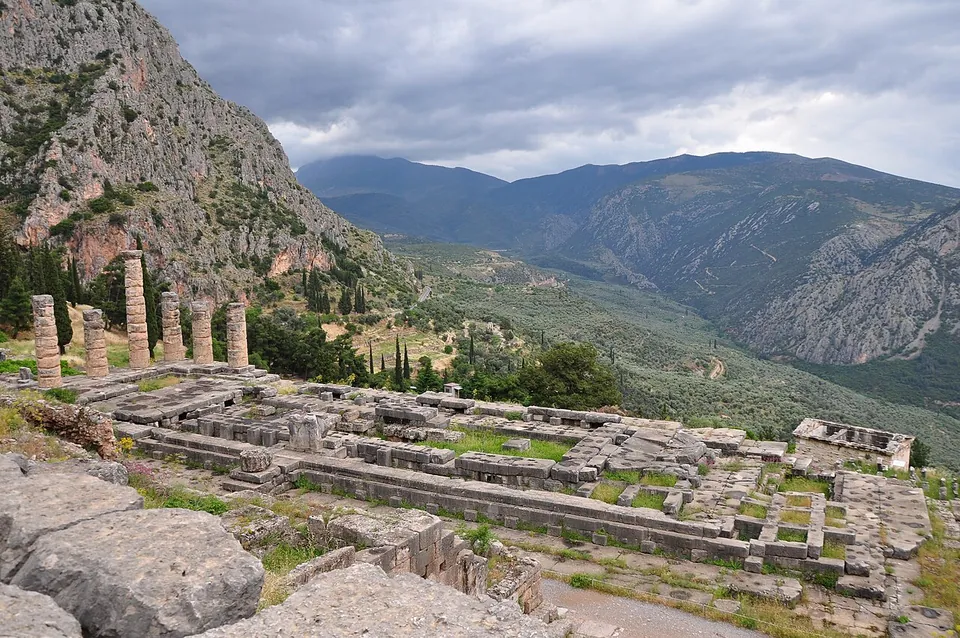 The long stone platform and foundations of a temple on a terrace, with a few columns standing at one end, below a steep rocky mountainside and above a deep green valley under a heavy grey sky.