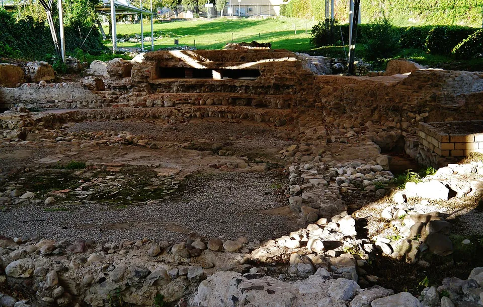 An excavated area under a white canopy, its floor marked by worn patches of mosaic and gravel, bounded by low rubble walls, with a hedge and lawn behind.