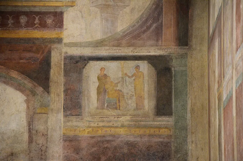 A section of painted wall in deep reds, ochres and greens imitating architecture, with a framed panel in the middle showing two standing figures and one seated.