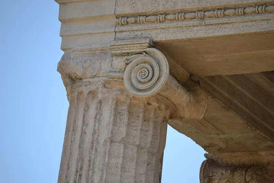 Close-up of the top of a fluted column with a spiral Ionic volute, under a moulded stone beam with a band of bead-and-reel ornament, against a blue sky.