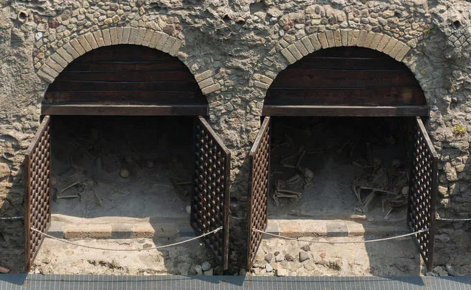 Two arched vaults built of rough stone with brick arches, behind open wooden lattice gates, their floors strewn with human skeletons.