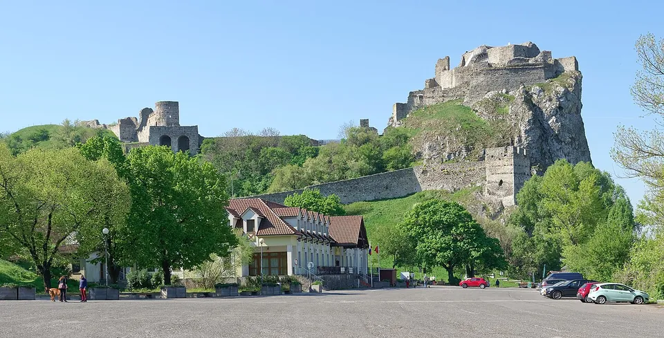 A paved square with parked cars and a restaurant building with a red roof below a wooded hill, on which the walls and towers of a castle climb to ruins on top of a tall rock.