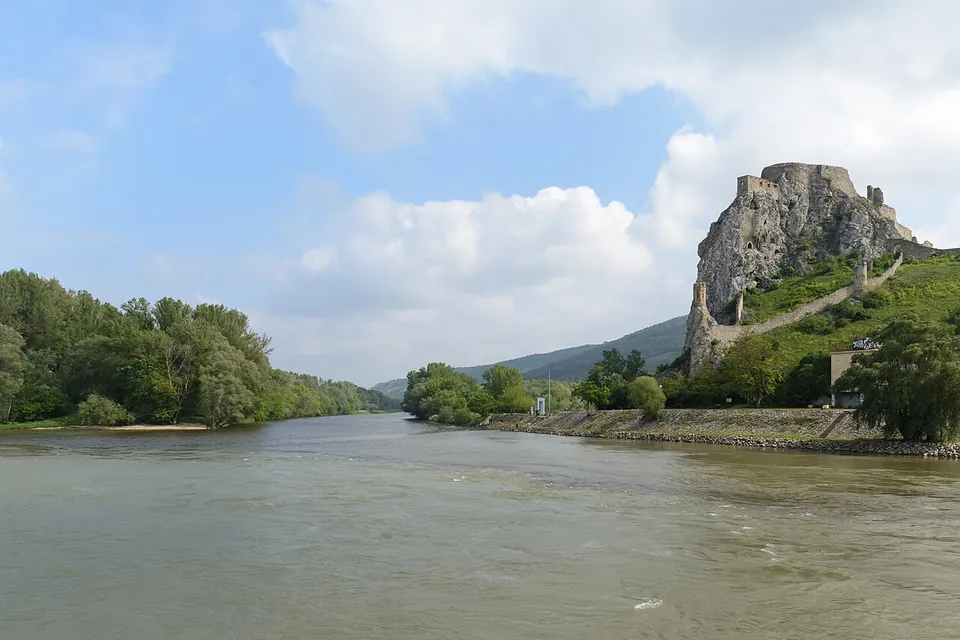A broad brown river in the foreground, with a tall grey rock on the right crowned by castle ruins and a long wall running down its grassy slope; wooded banks and a low hill fill the left.