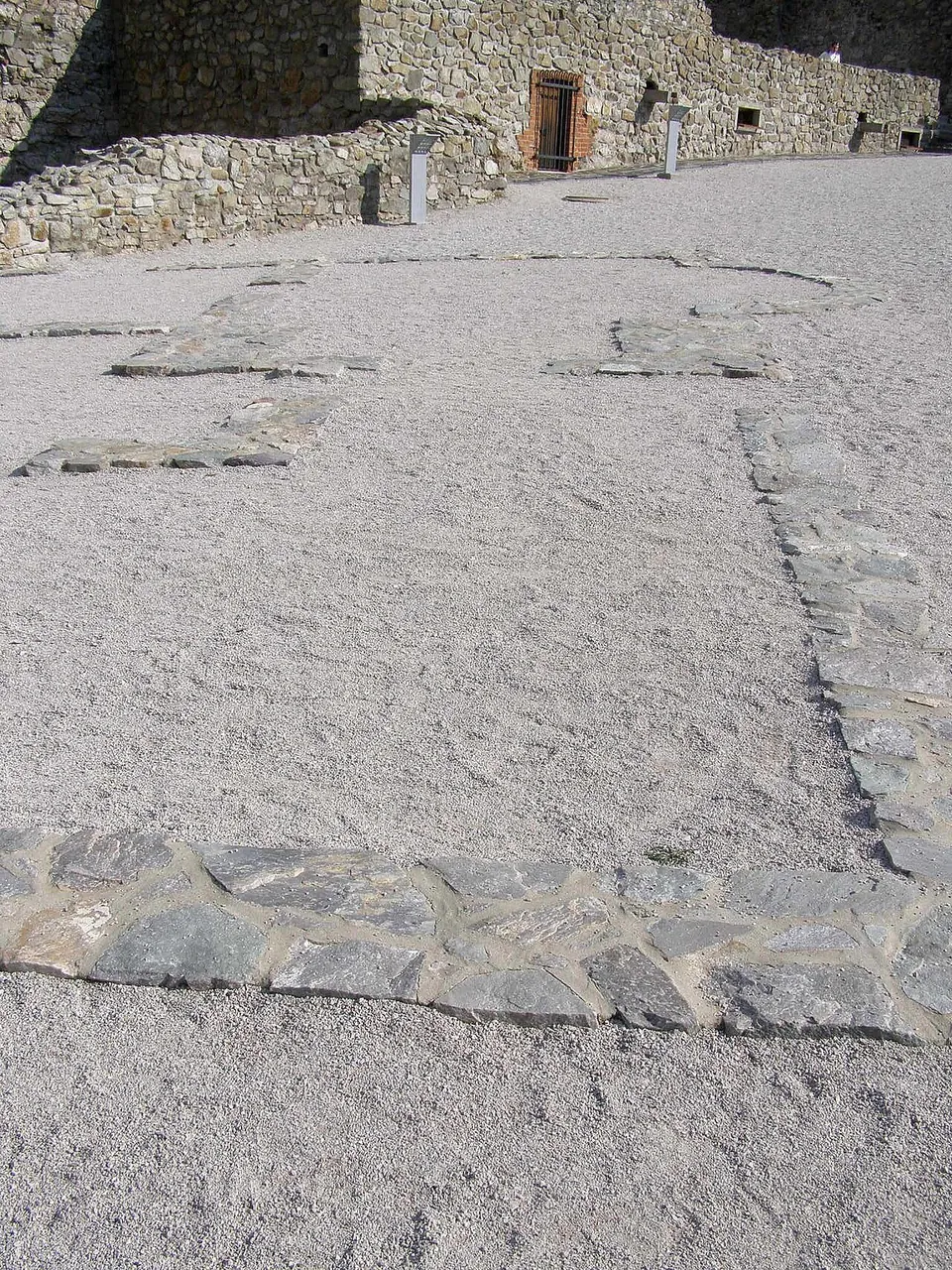 A gravel courtyard in which bands of flat grey stones laid at ground level trace the rectangular plan of a building, with a stretch of rubble castle wall, a small barred door and slim information posts behind.