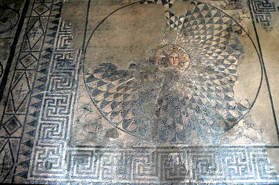A Roman floor mosaic with a small head of Medusa at the centre of a large circular shield of black and white scales, framed by a broad key-pattern border.