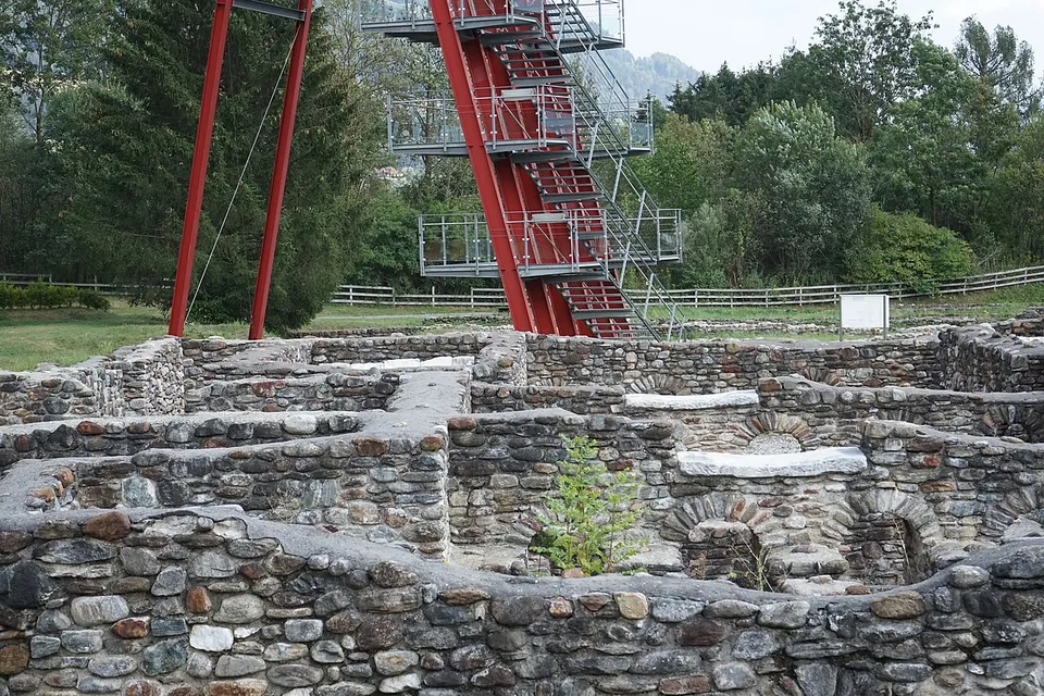 Close view of walls of rounded river stones set in mortar, a few courses high, with a red steel stair tower rising behind them among trees.