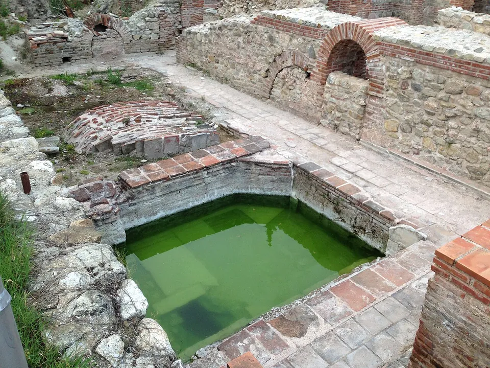 A rectangular pool of green water in the ruins of the baths, edged with brick and stone, with low walls, a small brick arch and paved paths around it.