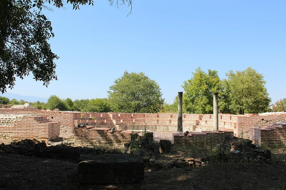 Restored low walls of red brick laid out in rows, with two standing columns among them and trees behind under a clear sky.