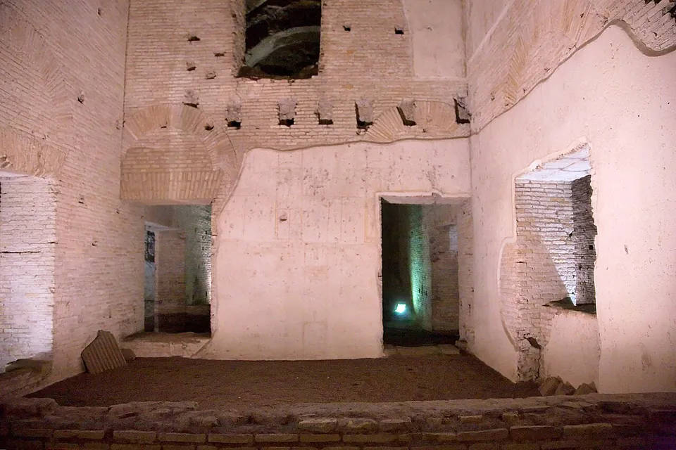 A lit underground room with high plastered and brick walls, doorways into further rooms, and the outline of a brick arch in the wall above.