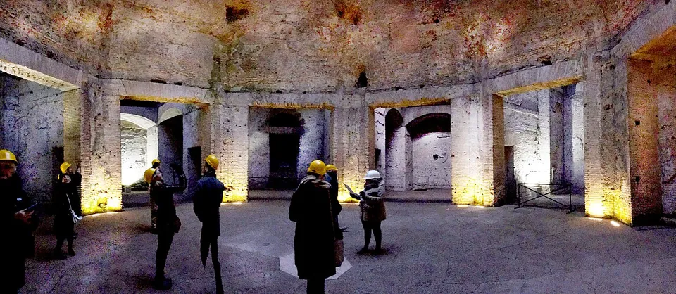 A wide view inside a dim, domed room of bare concrete and brick, lit from below, with tall square openings leading off into side rooms and a small group of visitors in yellow hard hats standing on the floor.