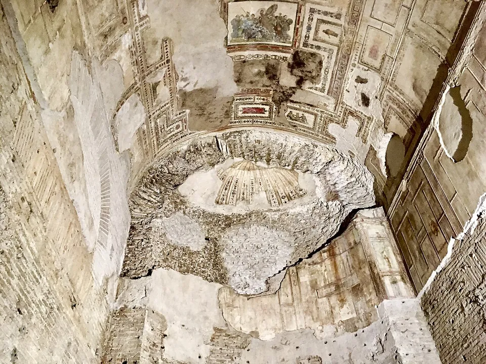 A vault painted with thin red and ochre frames around small figured panels, broken open around a shallow niche with a shell-shaped top, the plaster missing in patches down the walls.