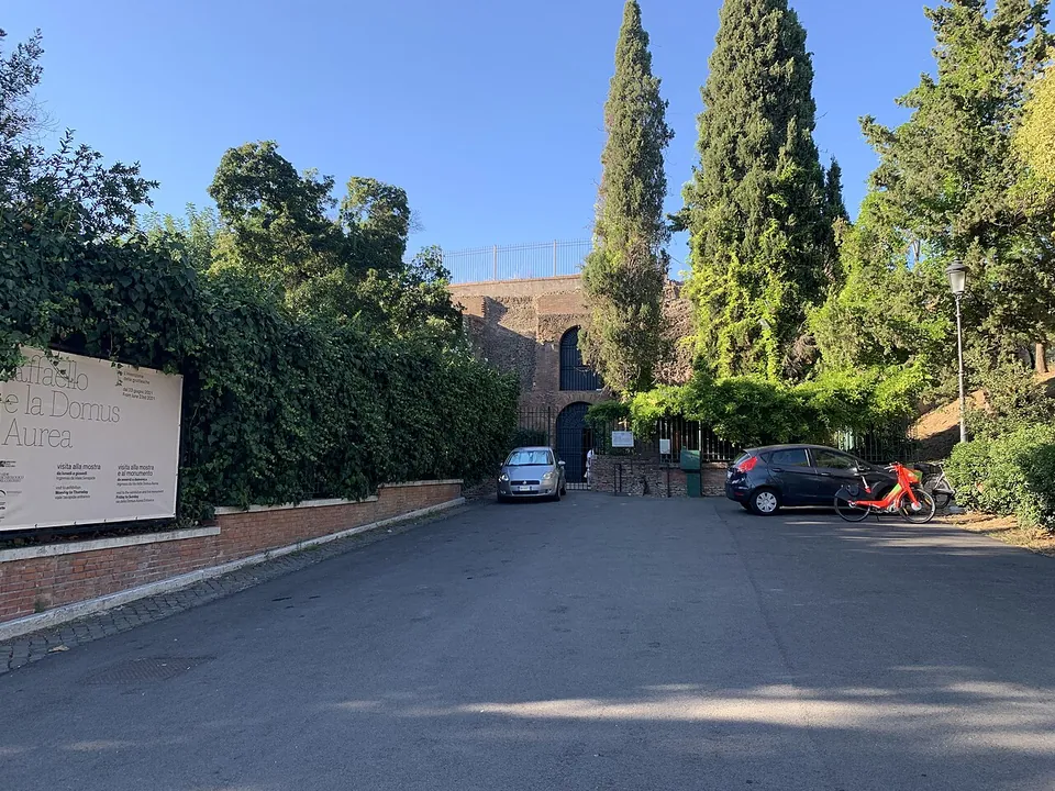 A short paved drive between hedges and cypresses, with a large sign naming the Domus Aurea on the left and a brick wall with an arched gate at the end, a few cars parked in front of it.