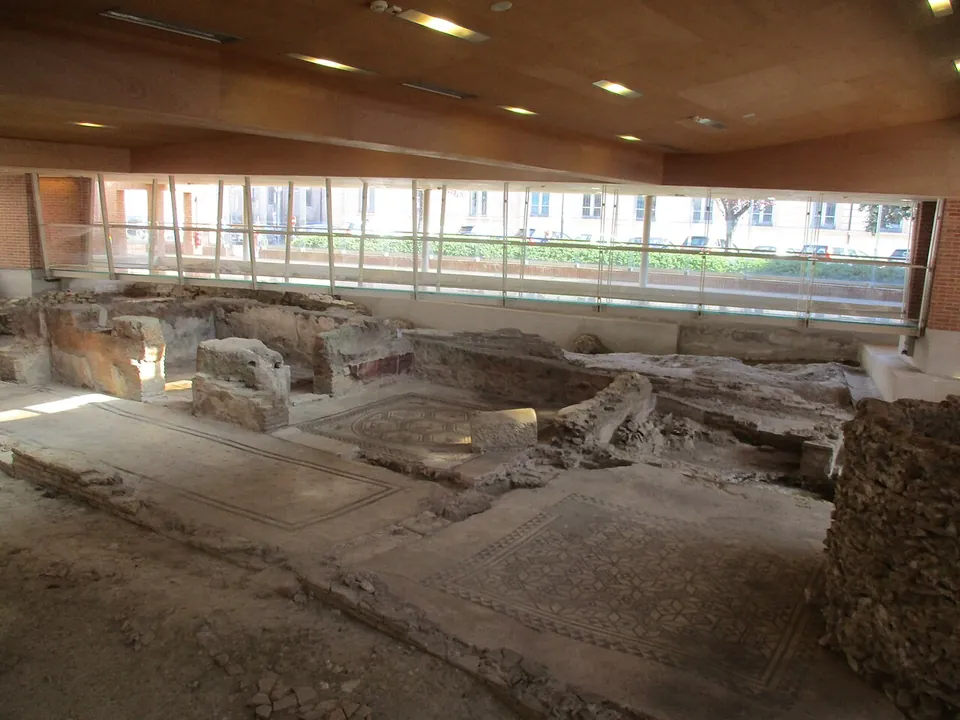 Inside a long modern hall with a wooden ceiling and a glass wall looking out onto a street, the low walls and patterned mosaic floors of excavated rooms lie below the visitors' level.