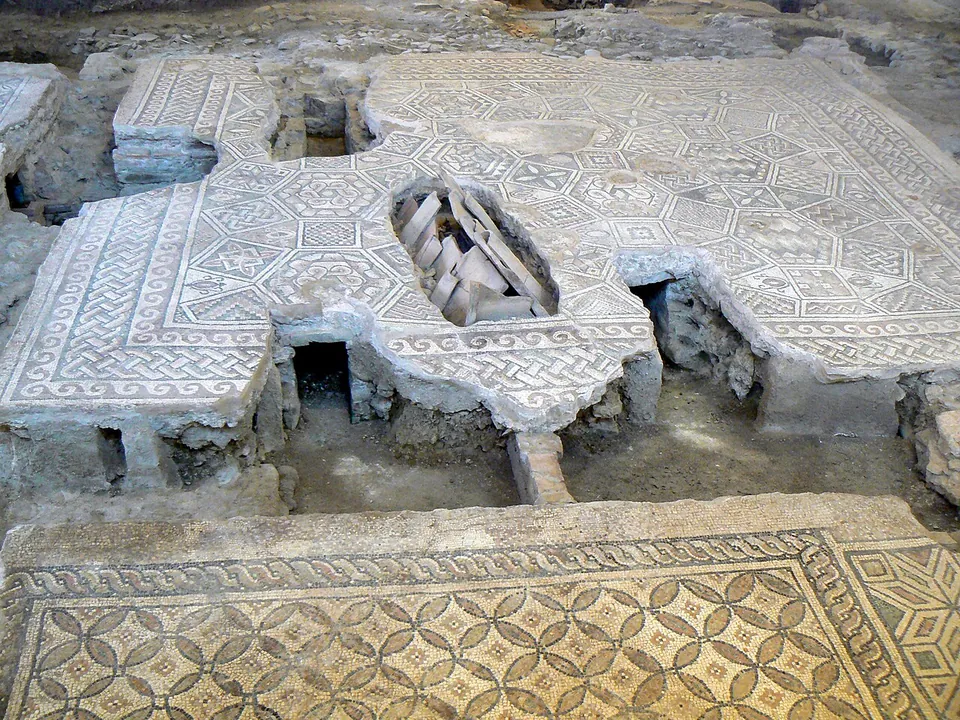 A white mosaic floor with a fine geometric pattern, broken through in places so that the low supports and the empty space beneath it show; another patterned floor lies in the foreground.