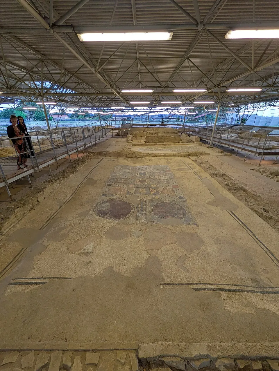 A long view down the inside of the shelter, with visitors standing on a railed walkway at the left and a run of mosaic floors with round patterned panels stretching away between low walls.