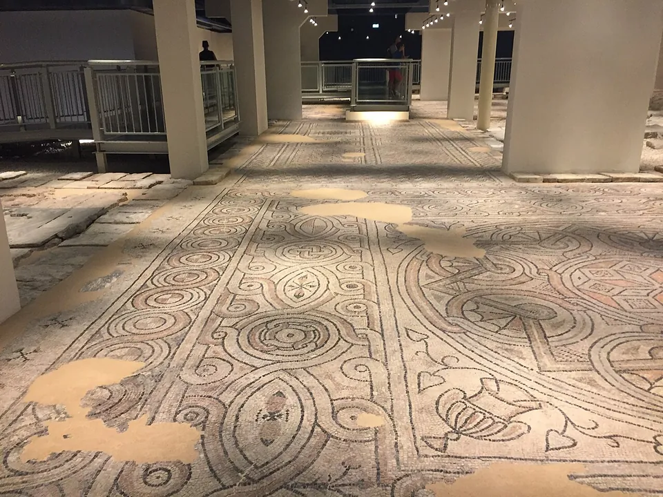 A mosaic floor of interlocking circles and ovals with small leaf and vase motifs, patched in places with plain mortar, running between white pillars to a railed walkway at the back.