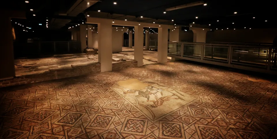A wide, low underground hall lit by ceiling spotlights, its floor covered with mosaic in a pattern of red and white squares, a figured panel at the centre, square white pillars carrying the roof and a glass-sided walkway along the right.