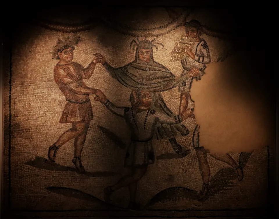 A dimly lit floor mosaic of four figures in short tunics dancing hand in hand in a ring, one wearing a wreath of leaves; part of the mosaic is lost at the right.