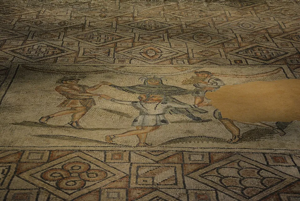 Seen at an angle on the floor, a figured mosaic panel of people in short tunics dancing hand in hand, part of it lost and filled with plain mortar, framed by a carpet of squares and lozenges with geometric and leaf motifs.