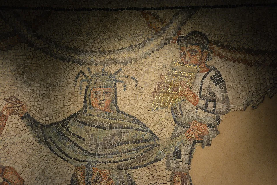 Close-up of mosaic tesserae showing a hooded figure wrapped in a dark green cloak and, beside it, a young man playing a set of pan pipes.