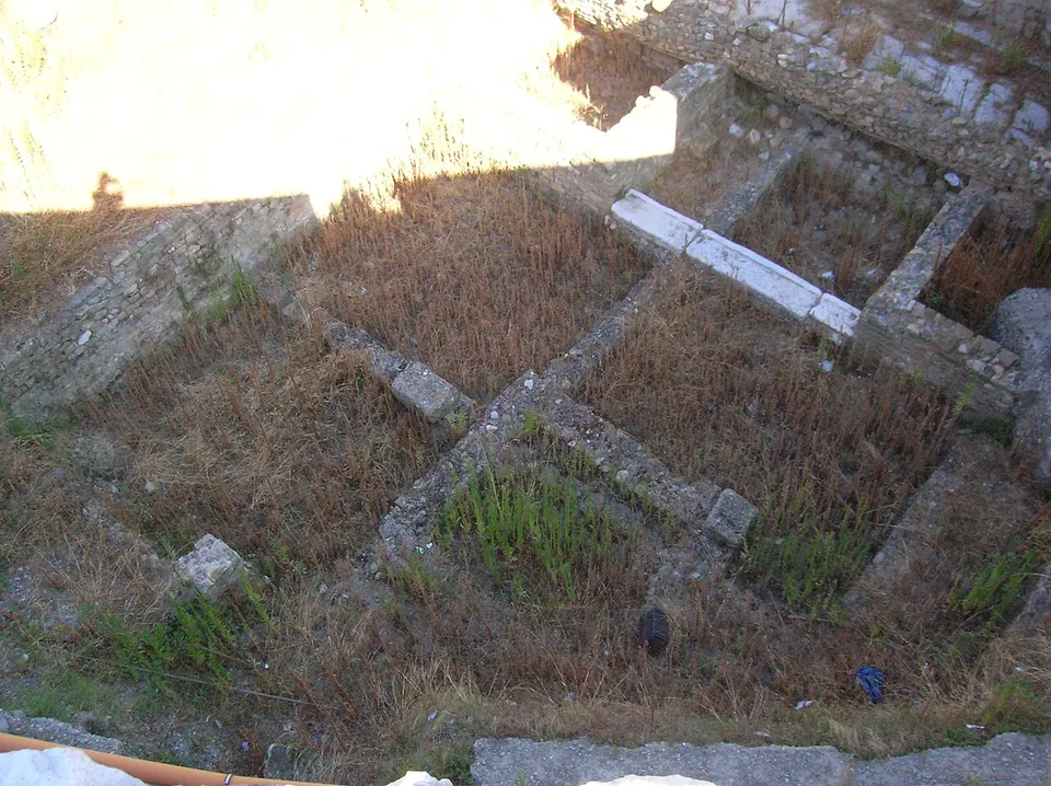 Looking down on the stone foundations of small rectangular rooms, overgrown with dry grass, with a line of white stone blocks along one wall.