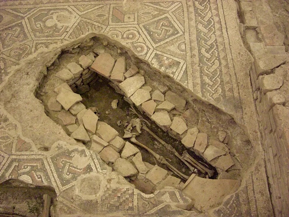 An oval grave lined with rough stones and broken tile, cut down through a multicoloured geometric mosaic floor, with bones visible at the bottom.