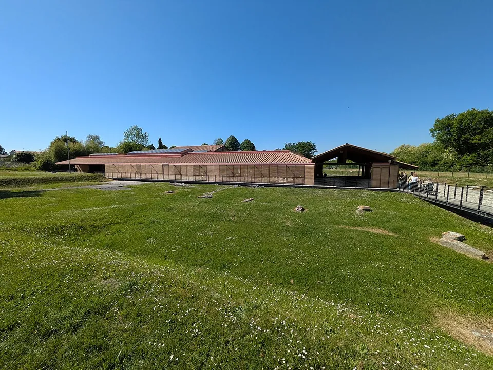 A long, low modern building with a red tiled roof and open sides, standing at the far end of a grassy field scattered with a few stones, with a raised walkway running along its side.