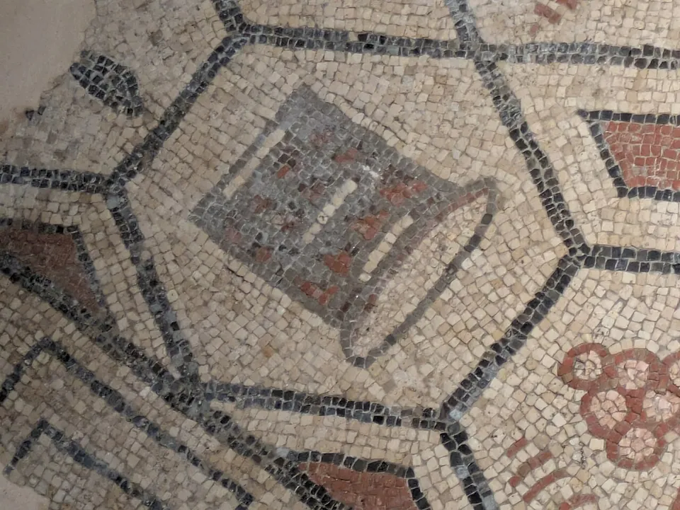 A close-up of a mosaic of small cream tesserae, divided by black lines into hexagons, one holding a grey and red motif and another a cluster of red rings.